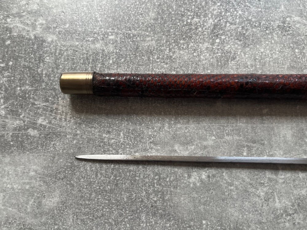 An antique French sword stick with a curved antler handle, brass collars, a narrow steel blade, and a sheath wrapped in dark brown snakeskin. View IV