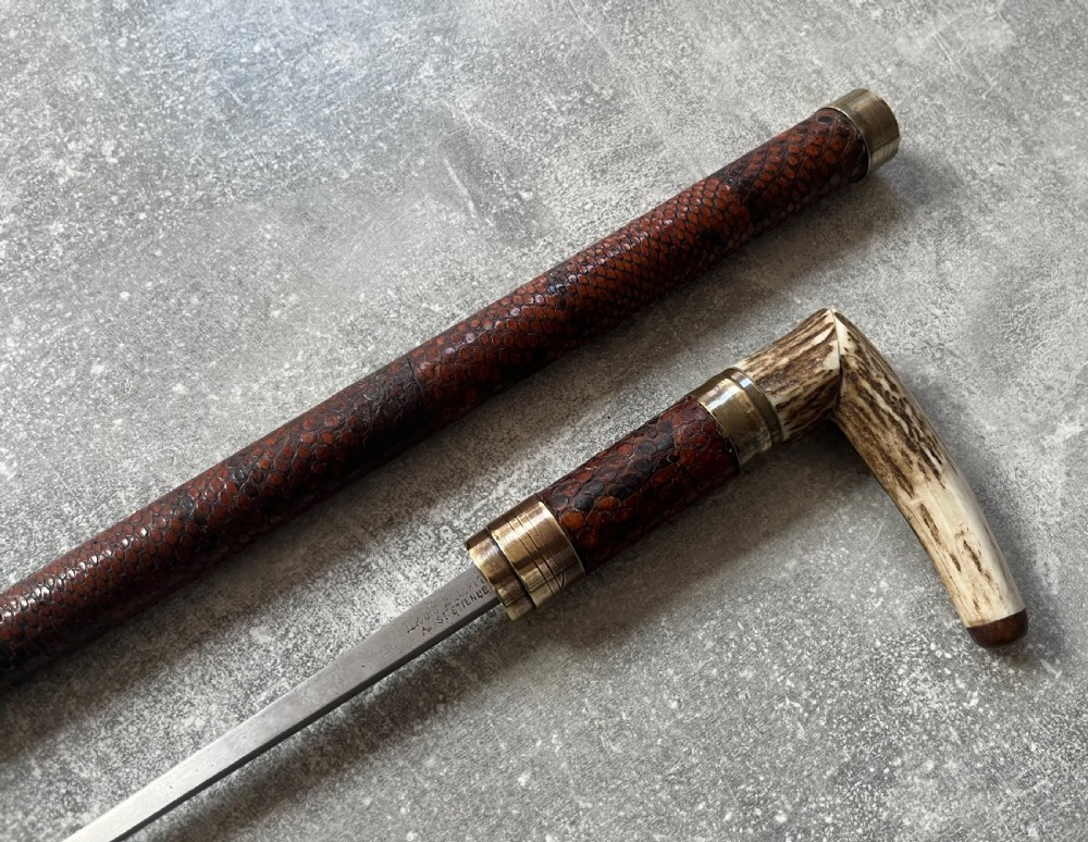 An antique French sword stick with a curved antler handle, brass collars, a narrow steel blade, and a sheath wrapped in dark brown snakeskin. View V