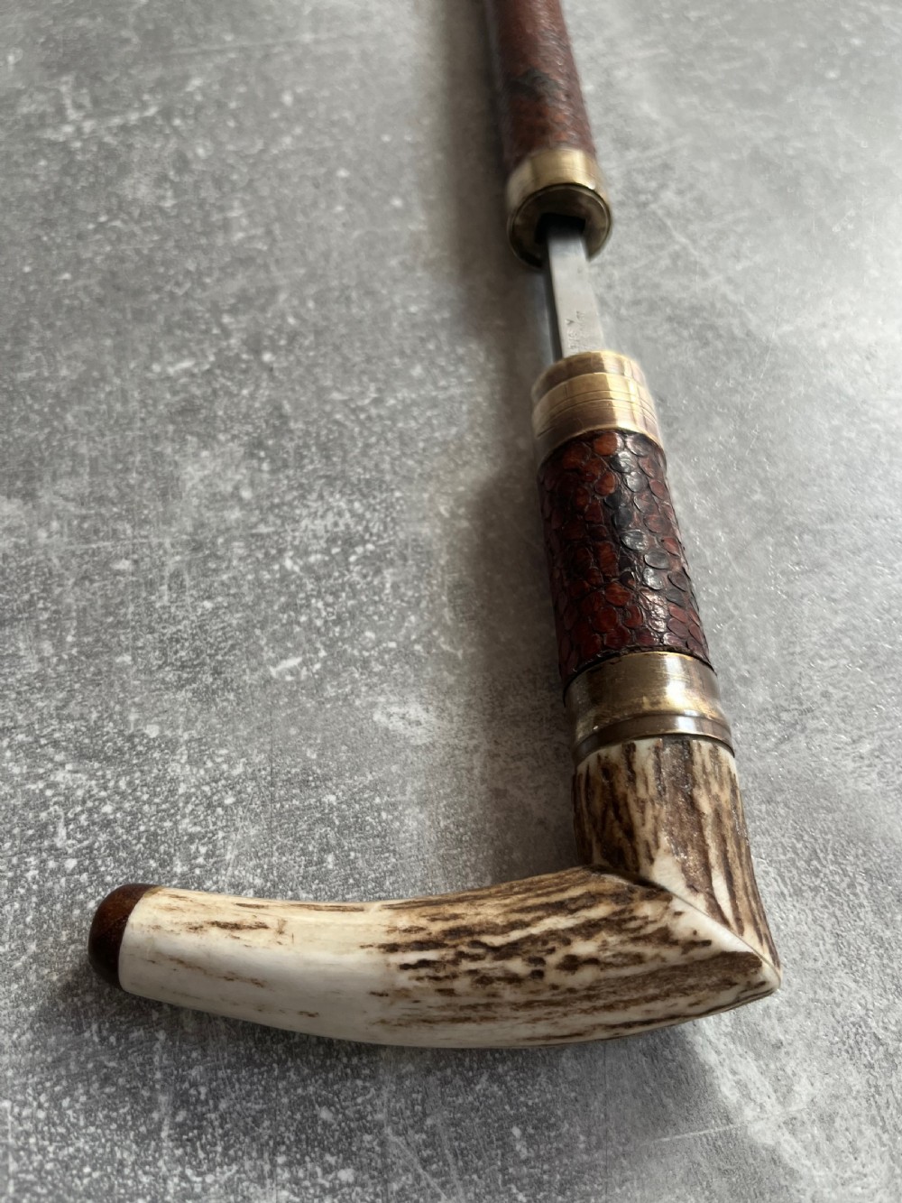 An antique French sword stick with a curved antler handle, brass collars, a narrow steel blade, and a sheath wrapped in dark brown snakeskin. View VI