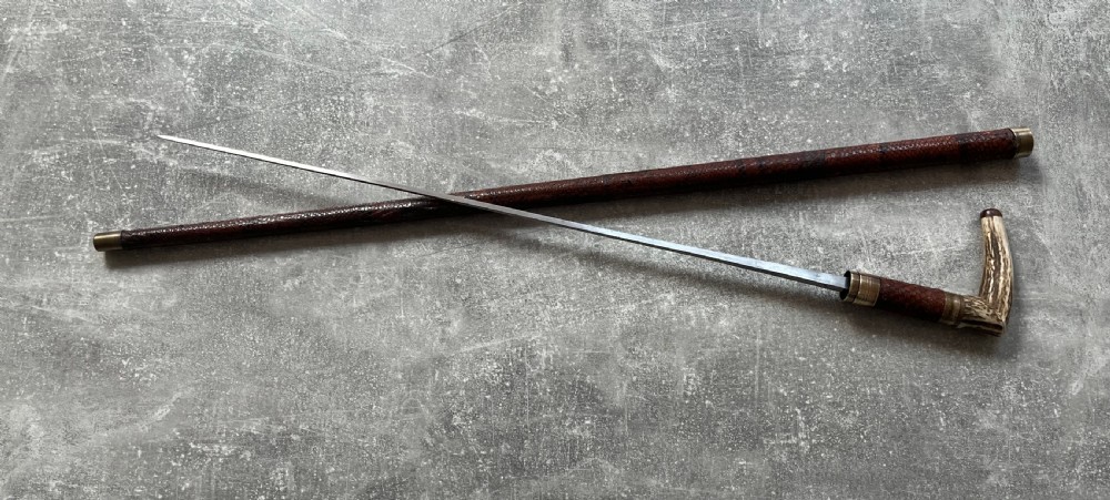 An antique French sword stick with a curved antler handle, brass collars, a narrow steel blade, and a sheath wrapped in dark brown snakeskin. View IX