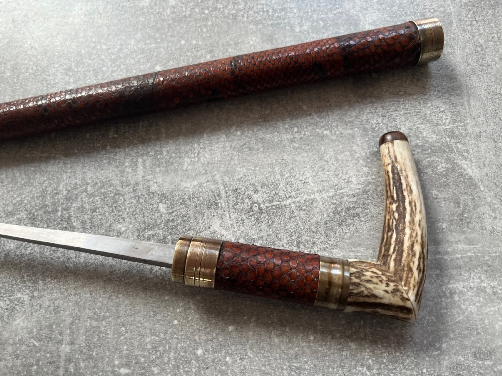 An antique French sword stick with a curved antler handle, brass collars, a narrow steel blade, and a sheath wrapped in dark brown snakeskin. View X