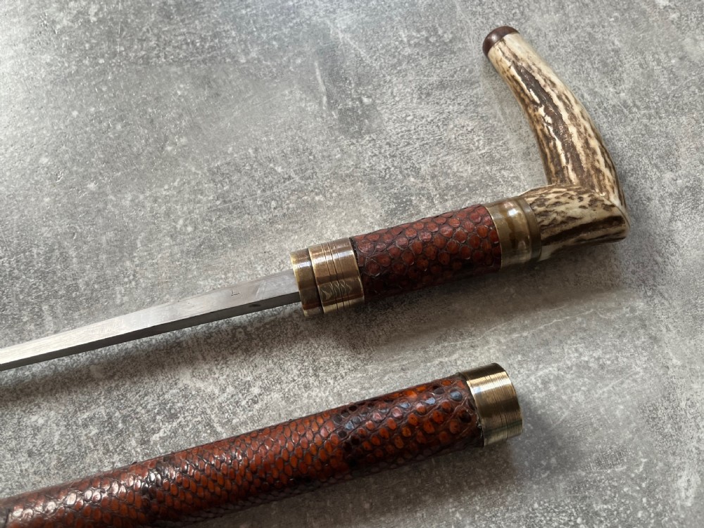 An antique French sword stick with a curved antler handle, brass collars, a narrow steel blade, and a sheath wrapped in dark brown snakeskin. View XI