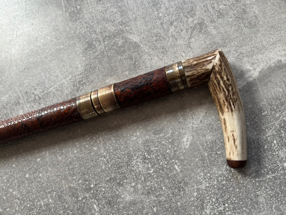An antique French sword stick with a curved antler handle, brass collars, a narrow steel blade, and a sheath wrapped in dark brown snakeskin. View XII