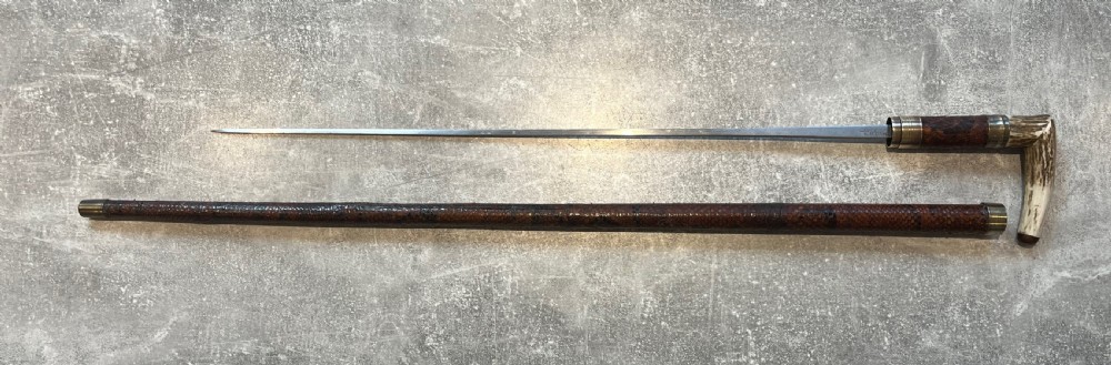An antique French sword stick with a curved antler handle, brass collars, a narrow steel blade, and a sheath wrapped in dark brown snakeskin. View XIV
