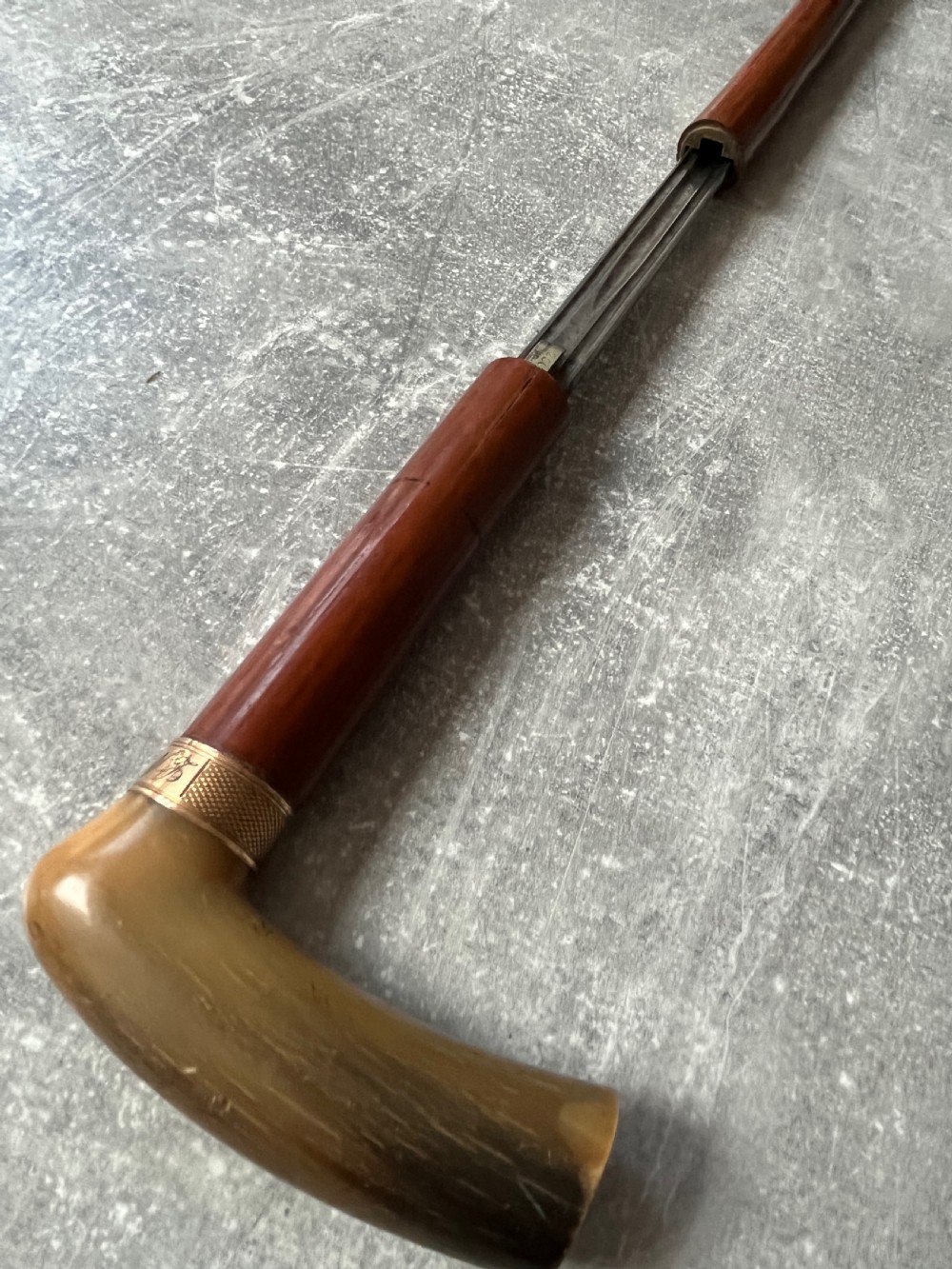 An antique French sword stick featuring an L-shaped horn handle, a gold-coloured engraved collar, a dark wooden sheath, and a slender unsheathed steel blade. View VI