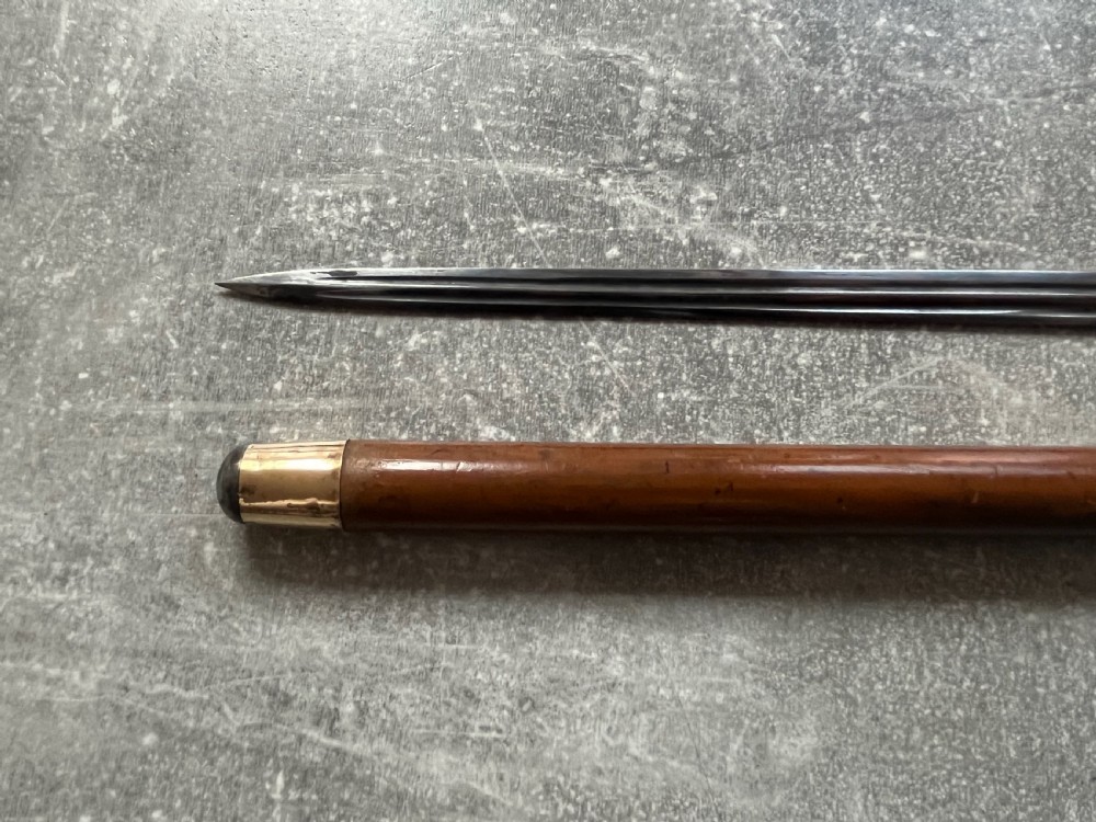 An antique French sword stick featuring an L-shaped horn handle, a gold-coloured engraved collar, a dark wooden sheath, and a slender unsheathed steel blade. View VII