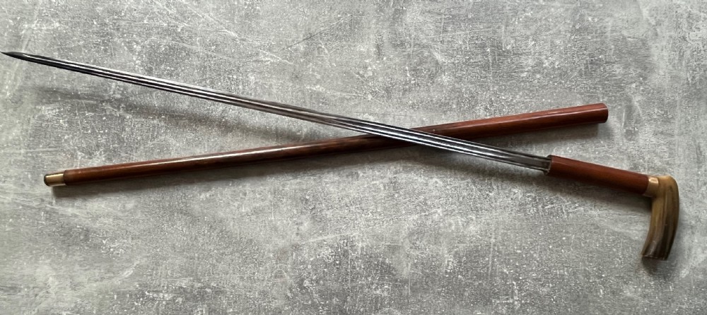 An antique French sword stick featuring an L-shaped horn handle, a gold-coloured engraved collar, a dark wooden sheath, and a slender unsheathed steel blade. View XI