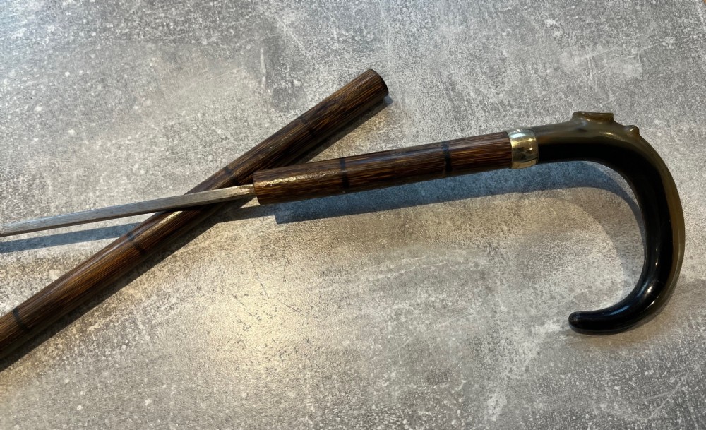 An antique Malacca walking stick unsheathed to reveal a long narrow steel sword blade, featuring a curved dark horn crook handle with a metal collar. View III