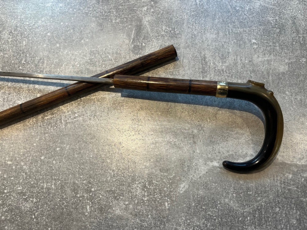 An antique Malacca walking stick unsheathed to reveal a long narrow steel sword blade, featuring a curved dark horn crook handle with a metal collar. View V