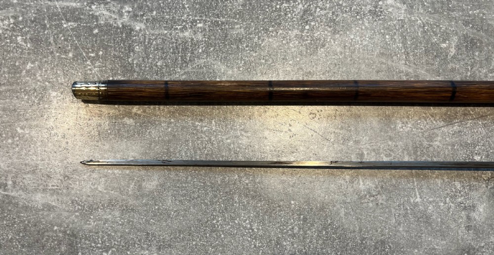 An antique Malacca walking stick unsheathed to reveal a long narrow steel sword blade, featuring a curved dark horn crook handle with a metal collar. View VI