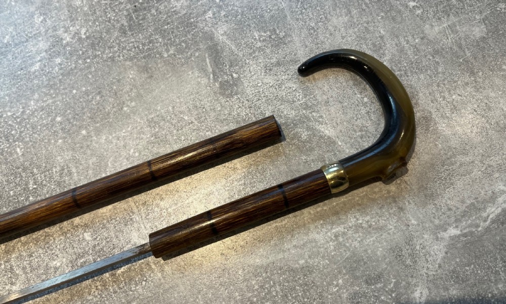 An antique Malacca walking stick unsheathed to reveal a long narrow steel sword blade, featuring a curved dark horn crook handle with a metal collar. View VII