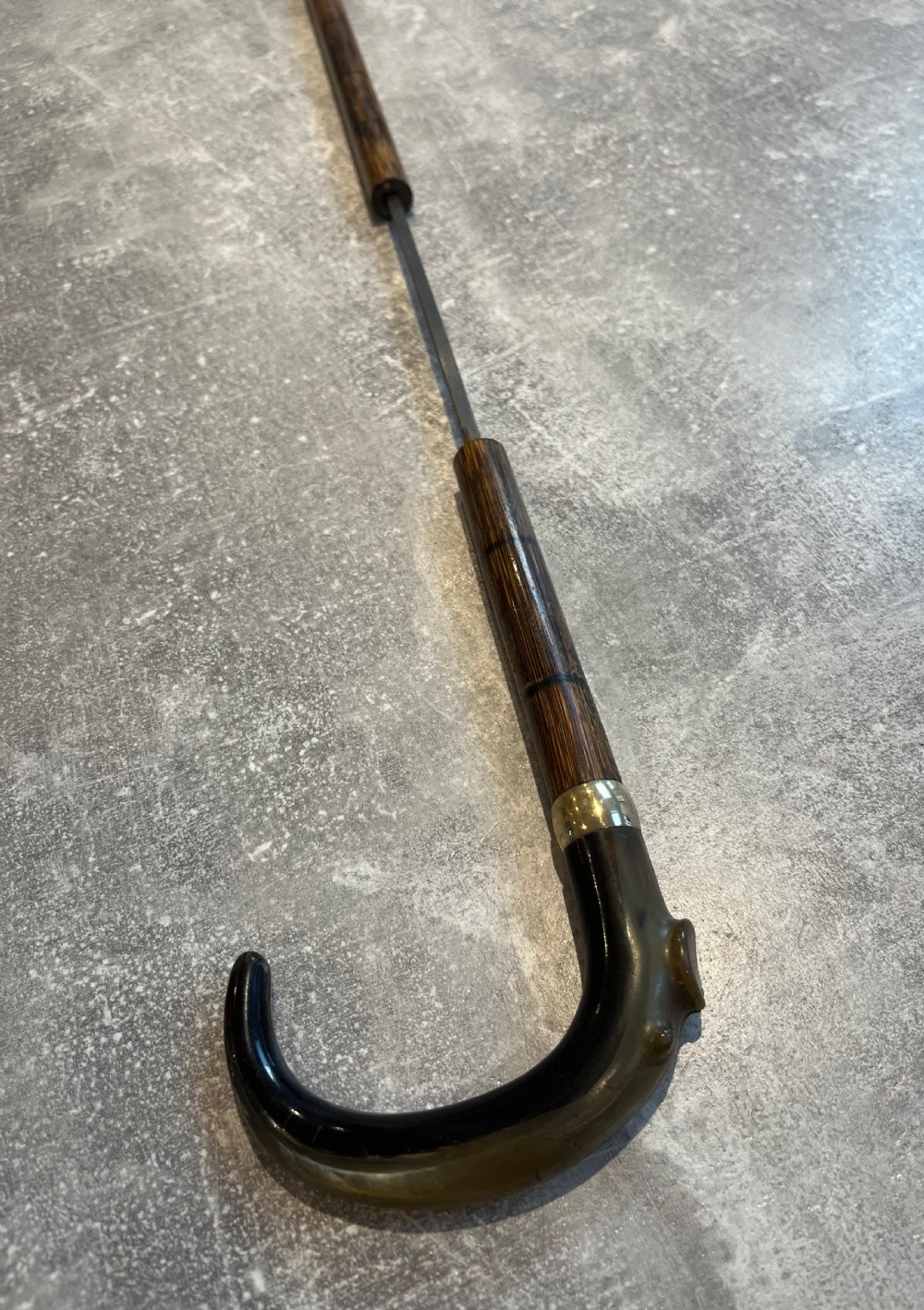An antique Malacca walking stick unsheathed to reveal a long narrow steel sword blade, featuring a curved dark horn crook handle with a metal collar. View IX