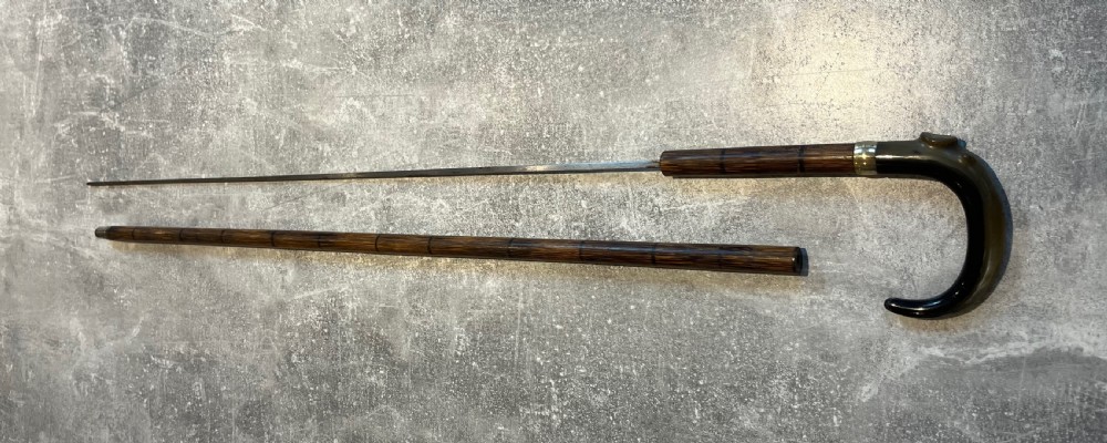 An antique Malacca walking stick unsheathed to reveal a long narrow steel sword blade, featuring a curved dark horn crook handle with a metal collar. View XV