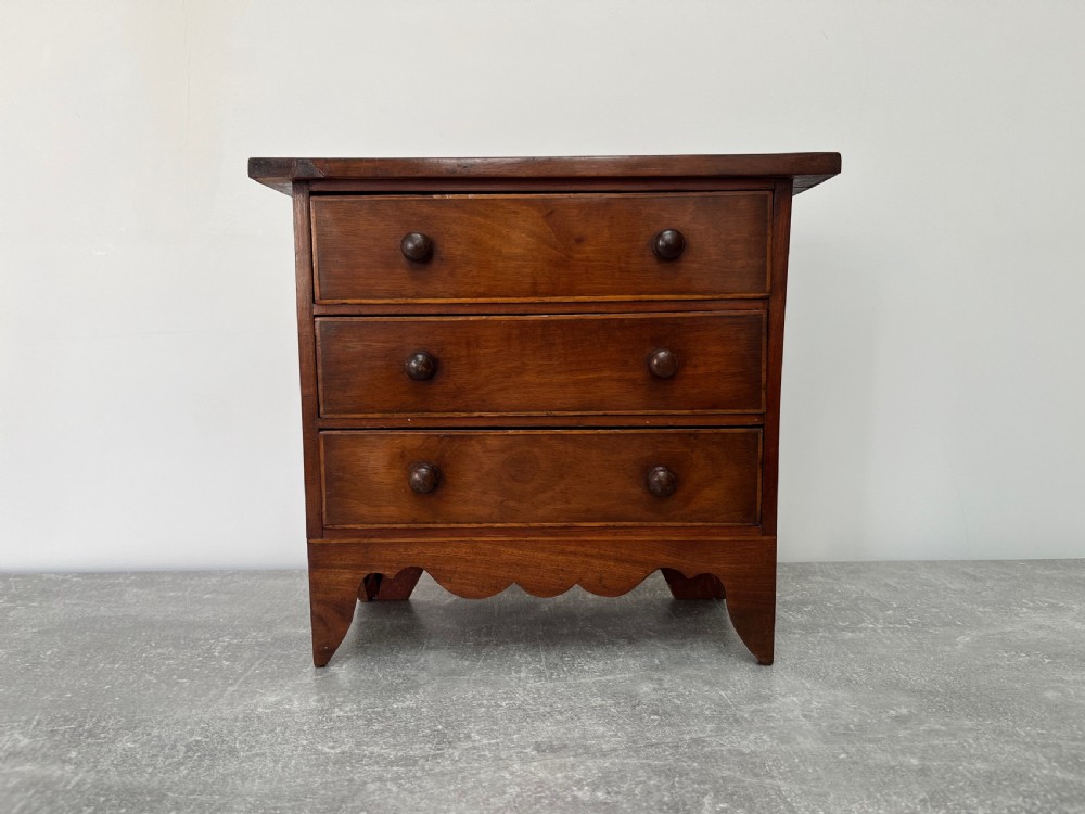 A miniature mahogany chest of drawers featuring three graduated drawers with turned wooden knobs and a shaped scalloped apron.