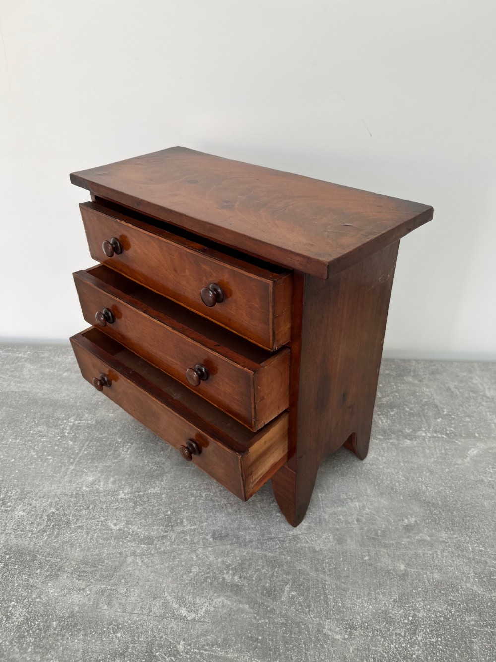 A miniature mahogany chest of drawers featuring three graduated drawers with turned wooden knobs and a shaped scalloped apron. View IV