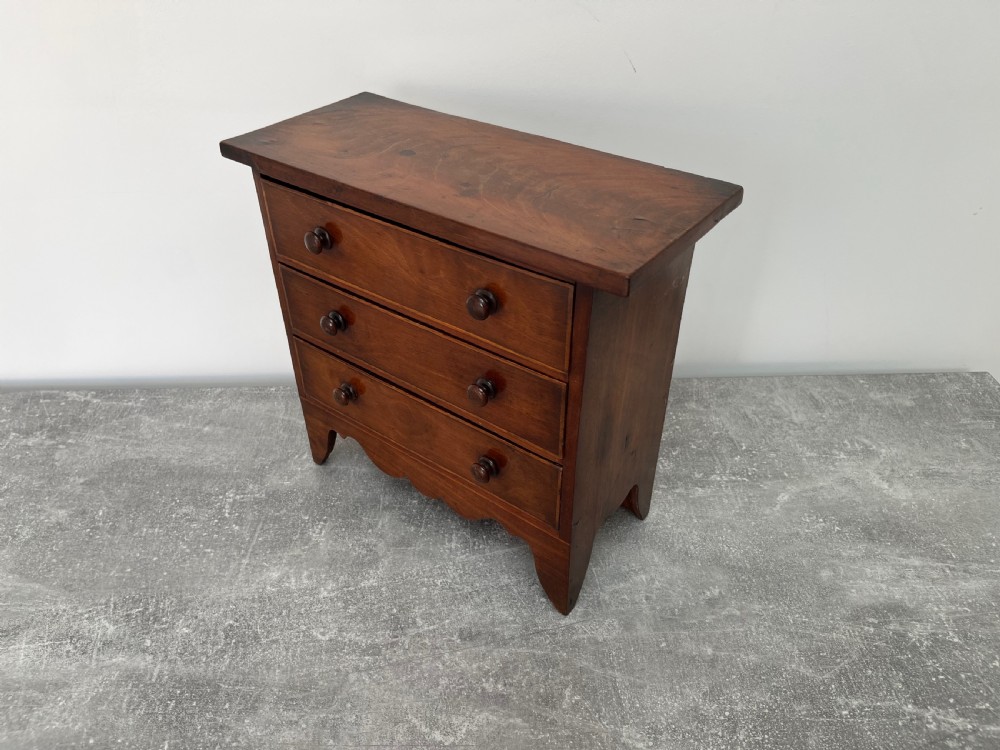 A miniature mahogany chest of drawers featuring three graduated drawers with turned wooden knobs and a shaped scalloped apron. View V