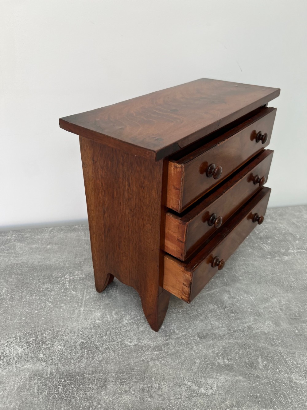 A miniature mahogany chest of drawers featuring three graduated drawers with turned wooden knobs and a shaped scalloped apron. View VI