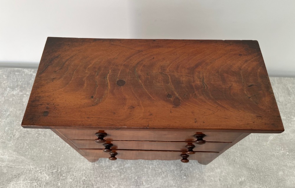 A miniature mahogany chest of drawers featuring three graduated drawers with turned wooden knobs and a shaped scalloped apron. View VIII