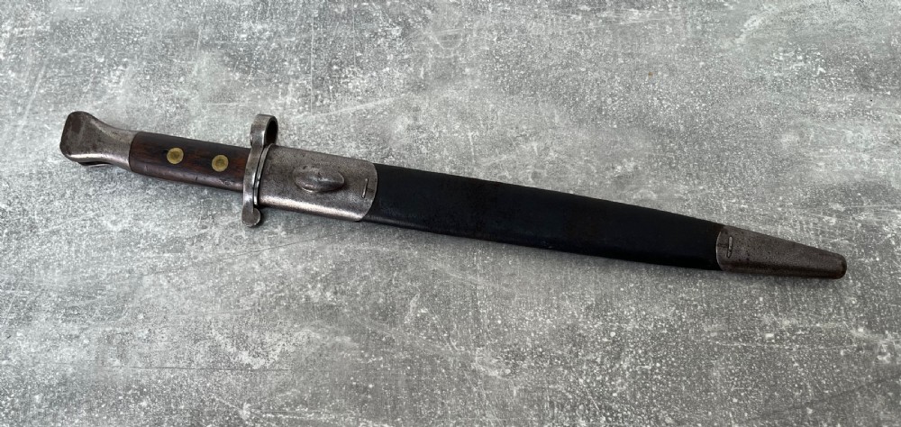 A Victorian British Pattern 1888 Mark 1 Type 2 sword bayonet with a double-edged steel blade, wooden handle grips secured with brass rivets, alongside its dark leather and steel-mounted scabbard. View III