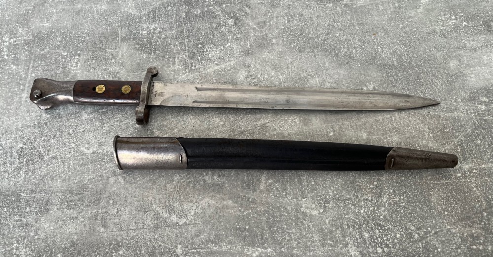 A Victorian British Pattern 1888 Mark 1 Type 2 sword bayonet with a double-edged steel blade, wooden handle grips secured with brass rivets, alongside its dark leather and steel-mounted scabbard. View IV