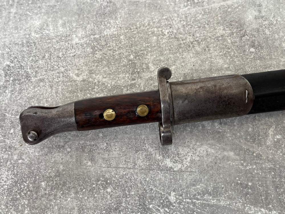 A Victorian British Pattern 1888 Mark 1 Type 2 sword bayonet with a double-edged steel blade, wooden handle grips secured with brass rivets, alongside its dark leather and steel-mounted scabbard. View IX
