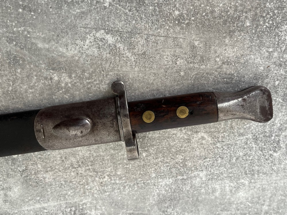 A Victorian British Pattern 1888 Mark 1 Type 2 sword bayonet with a double-edged steel blade, wooden handle grips secured with brass rivets, alongside its dark leather and steel-mounted scabbard. View XI