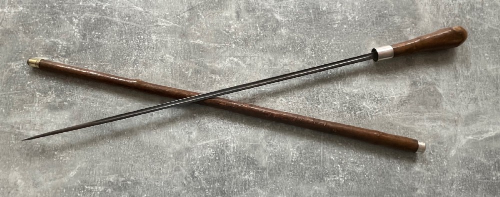 An antique chestnut wood sword stick shown unsheathed beside its shaft, featuring a root ball knob handle, silver collar mount, and long steel blade. View X