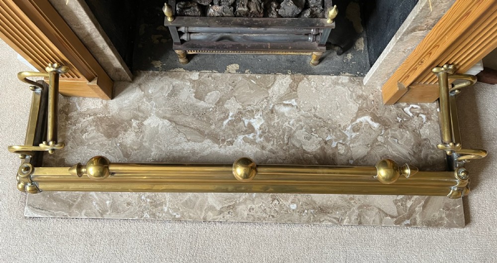 A low brass fire fender with rounded finials set around a hearth with matching brass companion tools on a small stand. View IX