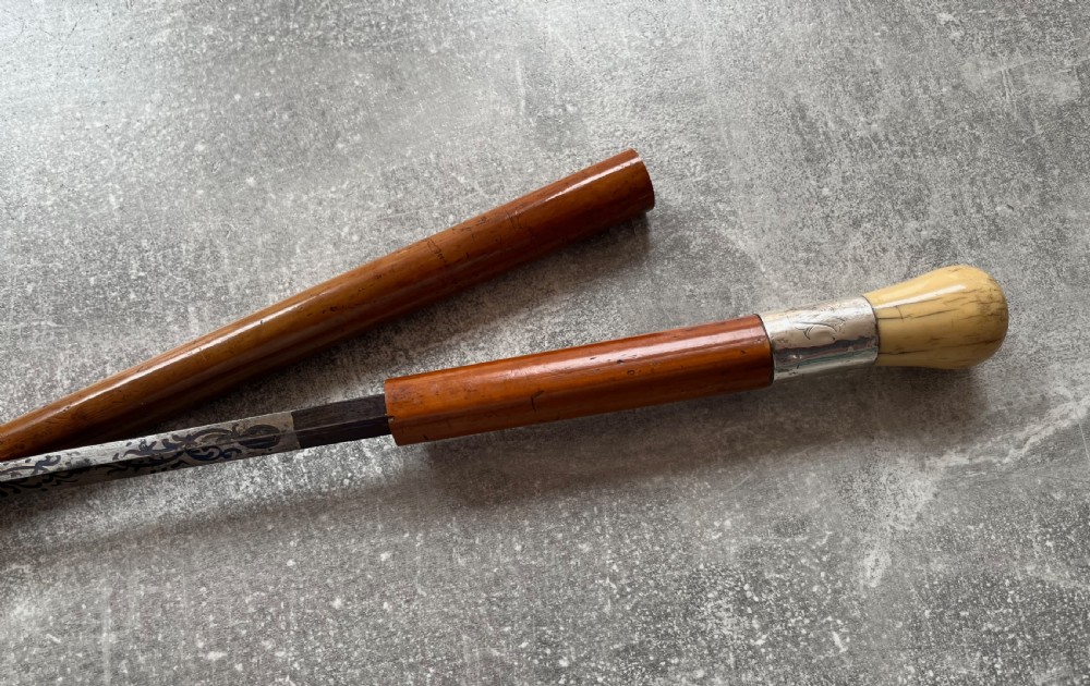 An antique Malacca sword stick shown unsheathed, featuring an etched steel blade, a polished wooden shaft, a silver collar, and a carved bone knop handle. View III