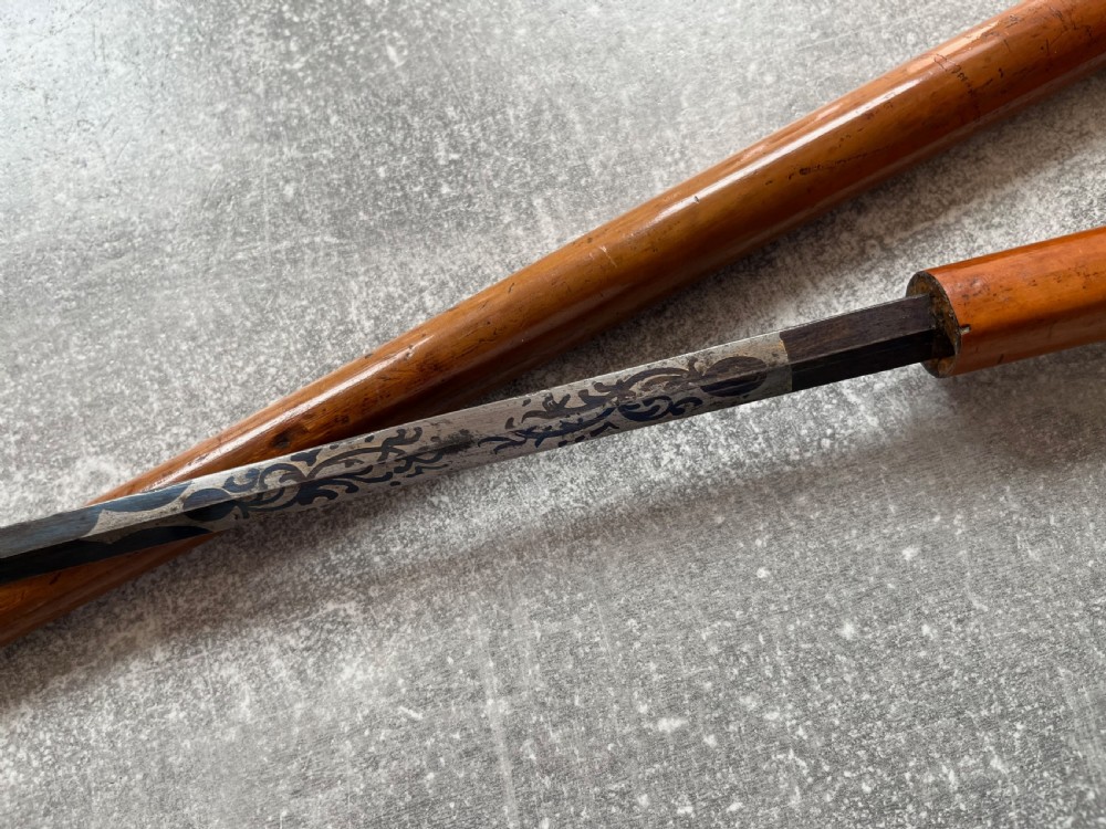 An antique Malacca sword stick shown unsheathed, featuring an etched steel blade, a polished wooden shaft, a silver collar, and a carved bone knop handle. View IV