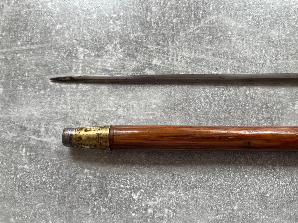 An antique Malacca sword stick shown unsheathed, featuring an etched steel blade, a polished wooden shaft, a silver collar, and a carved bone knop handle. View V