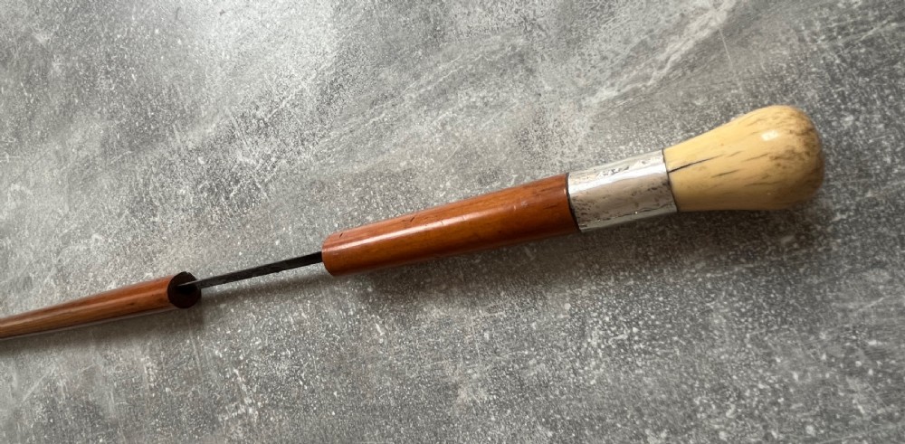 An antique Malacca sword stick shown unsheathed, featuring an etched steel blade, a polished wooden shaft, a silver collar, and a carved bone knop handle. View VIII