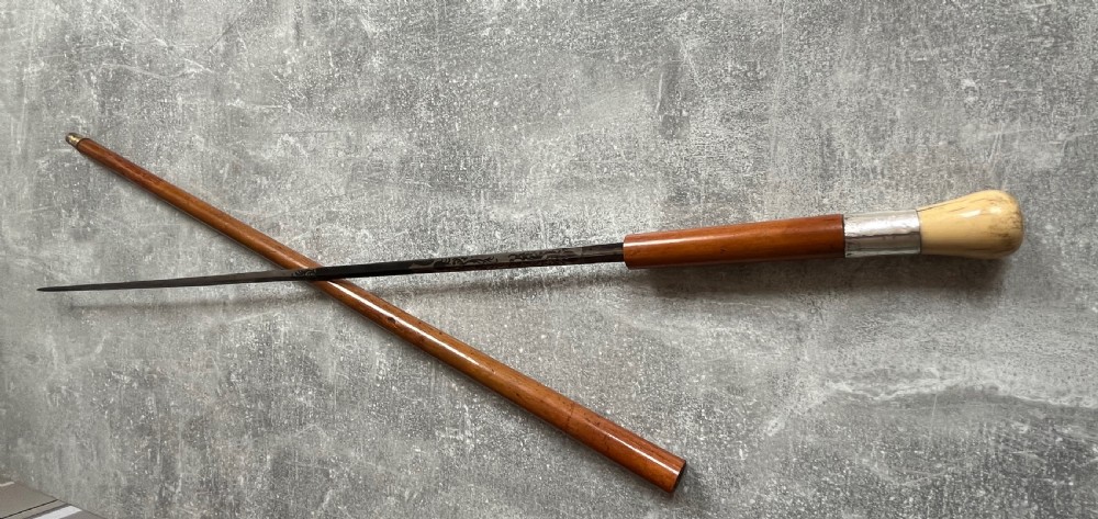 An antique Malacca sword stick shown unsheathed, featuring an etched steel blade, a polished wooden shaft, a silver collar, and a carved bone knop handle. View X
