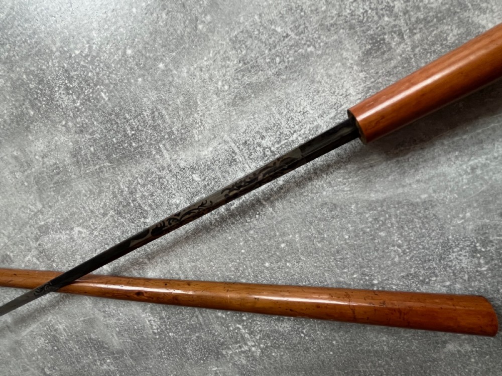 An antique Malacca sword stick shown unsheathed, featuring an etched steel blade, a polished wooden shaft, a silver collar, and a carved bone knop handle. View XI