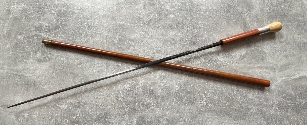 An antique Malacca sword stick shown unsheathed, featuring an etched steel blade, a polished wooden shaft, a silver collar, and a carved bone knop handle. View XII