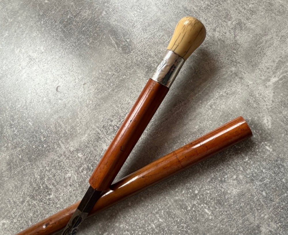 An antique Malacca sword stick shown unsheathed, featuring an etched steel blade, a polished wooden shaft, a silver collar, and a carved bone knop handle. View XIII