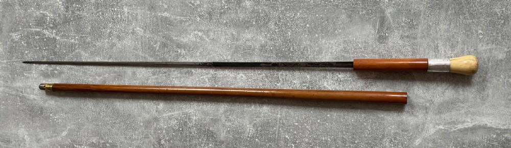 An antique Malacca sword stick shown unsheathed, featuring an etched steel blade, a polished wooden shaft, a silver collar, and a carved bone knop handle. View XIV