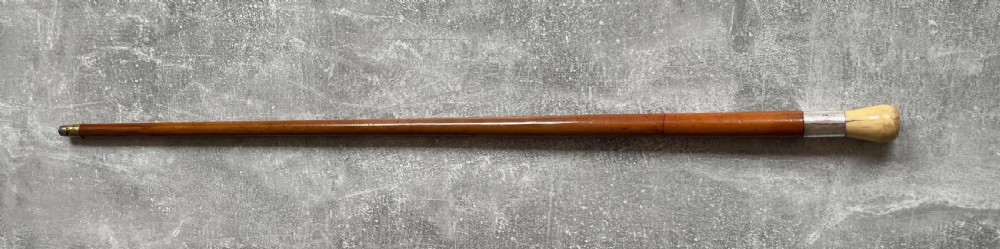 An antique Malacca sword stick shown unsheathed, featuring an etched steel blade, a polished wooden shaft, a silver collar, and a carved bone knop handle. View XV