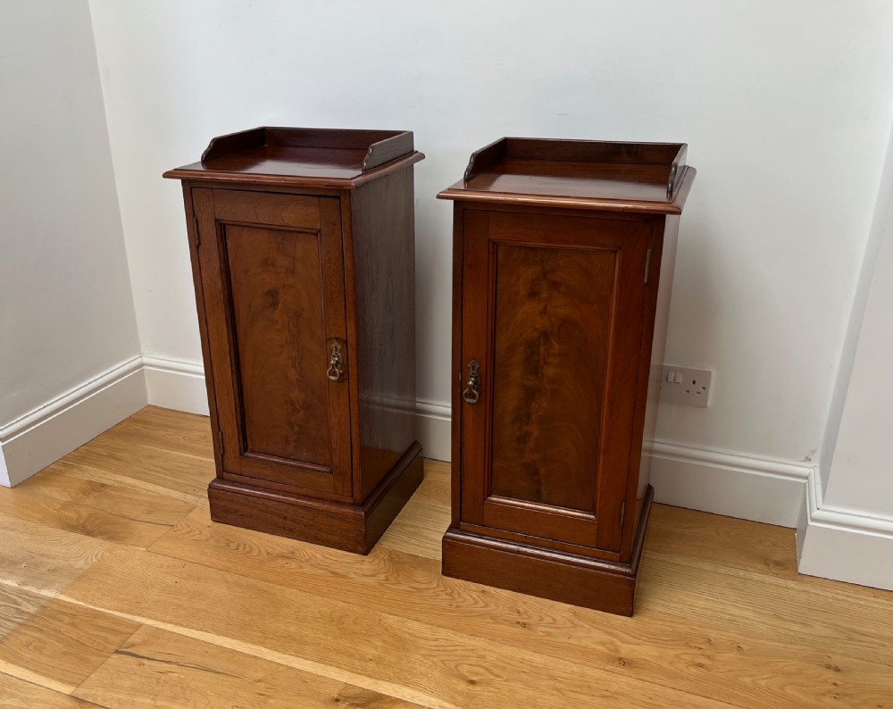 A pair of antique mahogany bedside cabinets featuring gallery tops, single panelled doors, brass drop handles, and solid plinth bases.