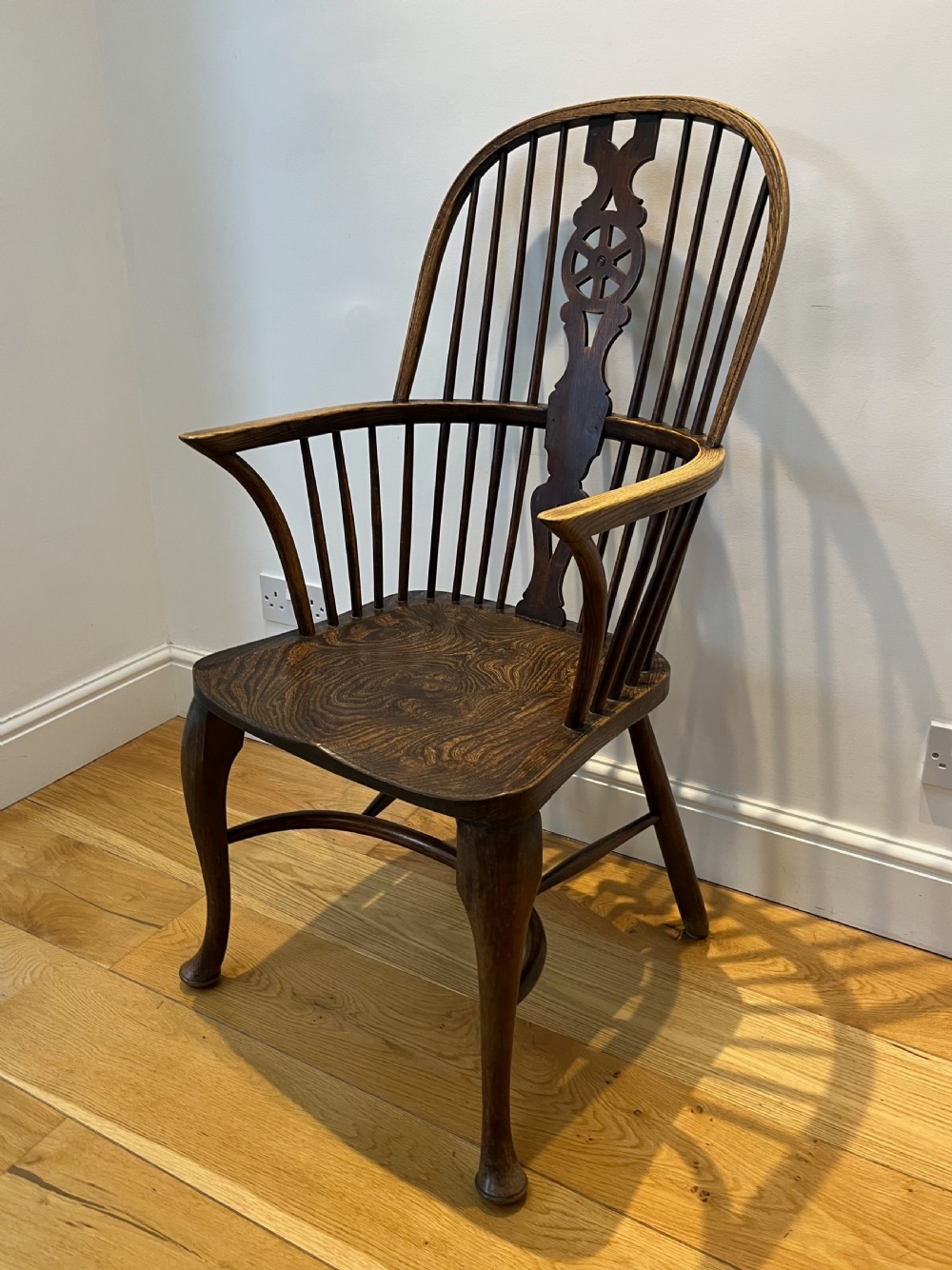 A dark brown wooden Windsor armchair featuring a high hoop back with a wheel-motif central splat, curved armrests, a contoured saddle seat, and front cabriole legs.