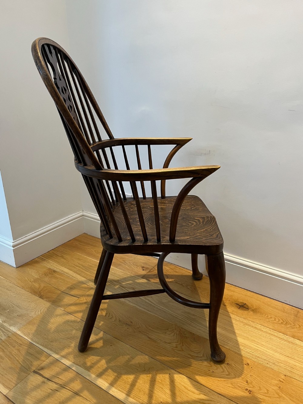 A dark brown wooden Windsor armchair featuring a high hoop back with a wheel-motif central splat, curved armrests, a contoured saddle seat, and front cabriole legs. View III