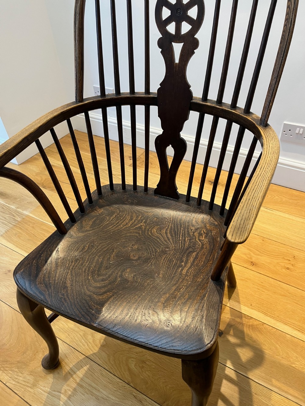 A dark brown wooden Windsor armchair featuring a high hoop back with a wheel-motif central splat, curved armrests, a contoured saddle seat, and front cabriole legs. View VI