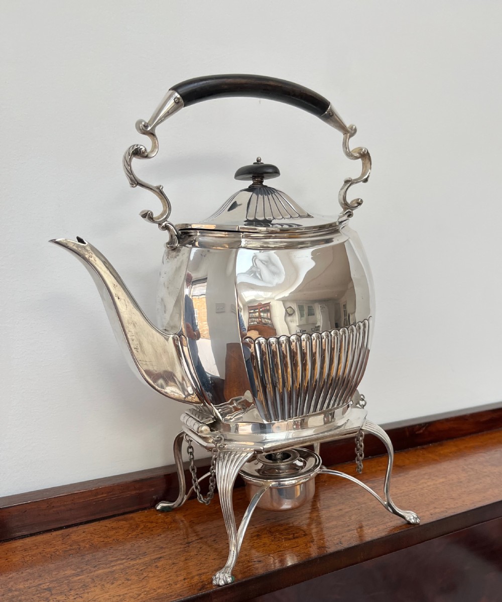 A silver-plated spirit kettle with a fluted lower body, dark wooden handle, gooseneck spout, and matching stand containing an alcohol burner below. View III