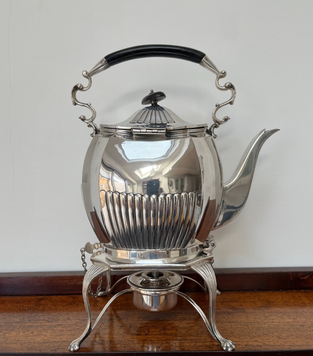 A silver-plated spirit kettle with a fluted lower body, dark wooden handle, gooseneck spout, and matching stand containing an alcohol burner below. View V