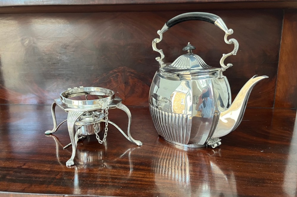 A silver-plated spirit kettle with a fluted lower body, dark wooden handle, gooseneck spout, and matching stand containing an alcohol burner below. View IX