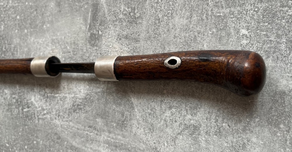 A late 19th-century dark hazel wood walking stick with a curved handle, silver collar, upper eyelet detail, and a metallic ferrule base. View X