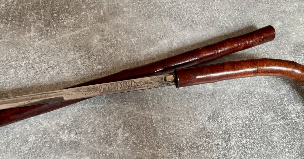 An antique cherrywood sword stick featuring a curved crook handle, a dark polished wood shaft, and a drawn steel blade with etched markings. View V