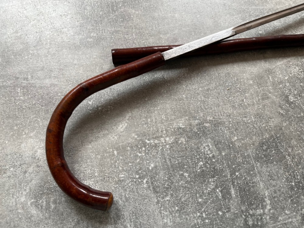 An antique cherrywood sword stick featuring a curved crook handle, a dark polished wood shaft, and a drawn steel blade with etched markings. View X