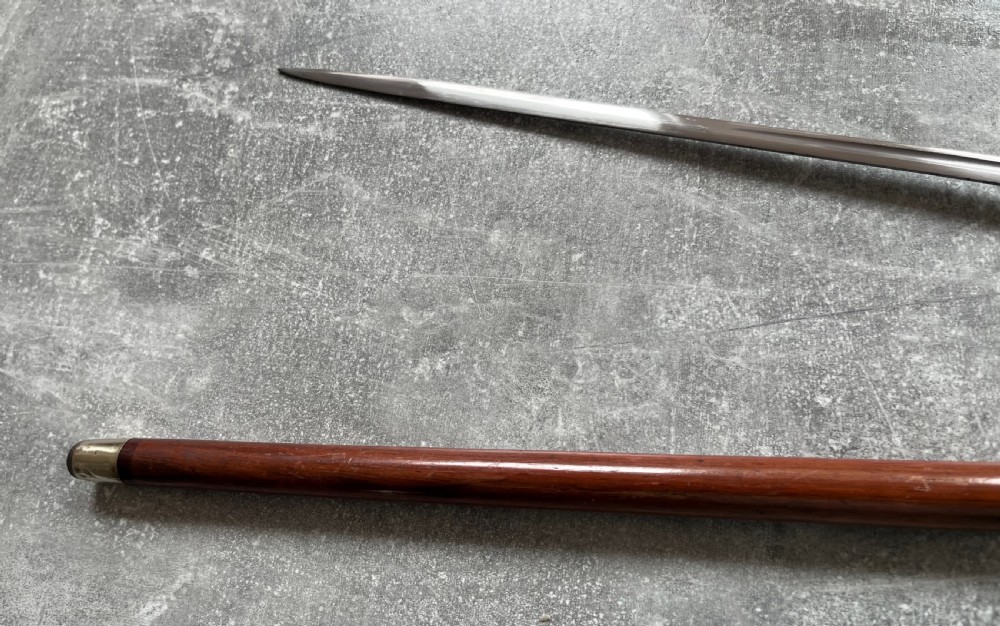 An antique brown Malacca wood walking stick with a curved crook handle, shown unsheathed to reveal an etched steel Toledo blade alongside its wooden sheath. View V