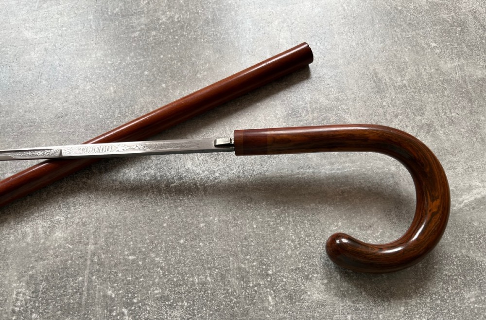 An antique brown Malacca wood walking stick with a curved crook handle, shown unsheathed to reveal an etched steel Toledo blade alongside its wooden sheath. View X